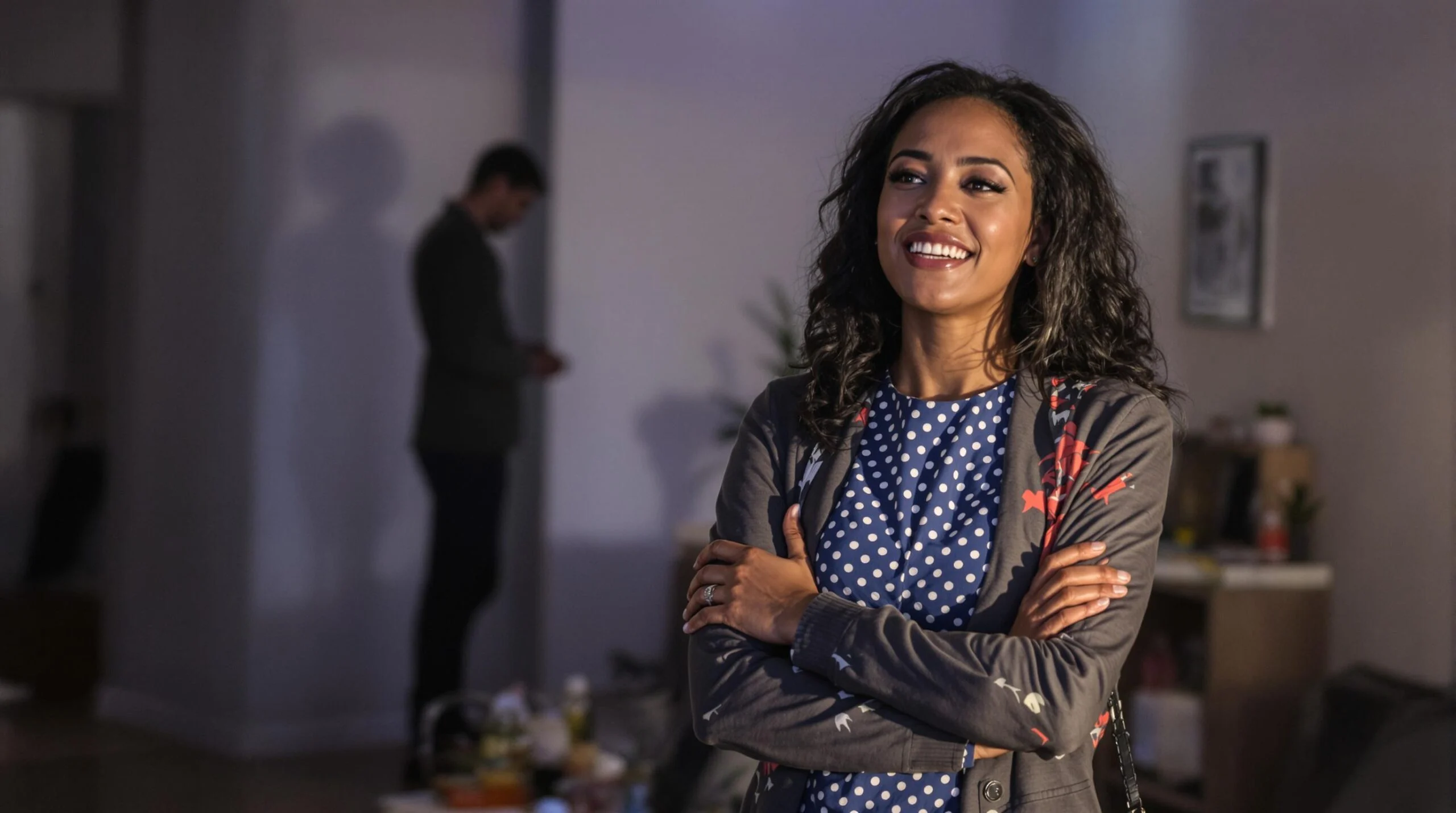 A confident woman smiling with her arms crossed, standing in a dimly lit room with someone blurred in the background.
