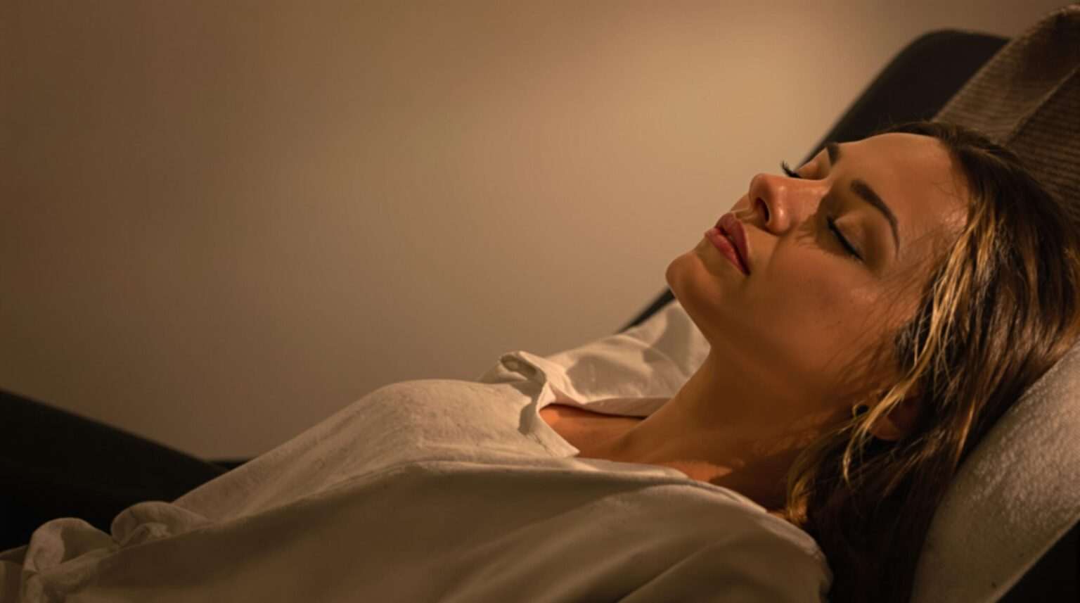 A relaxed woman lying down with her eyes closed, appearing calm in a dimly lit environment.