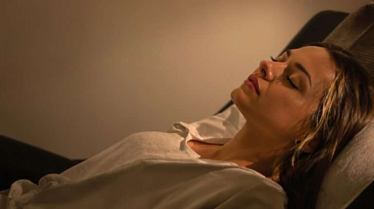 A relaxed woman lying down with her eyes closed, appearing calm in a dimly lit environment.