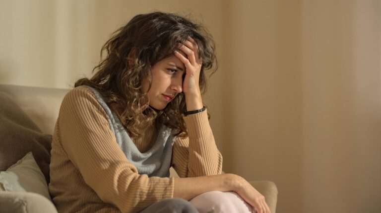 Young woman sitting on a couch with her hand on her forehead, appearing deep in thought and experiencing stress or anxiety.
