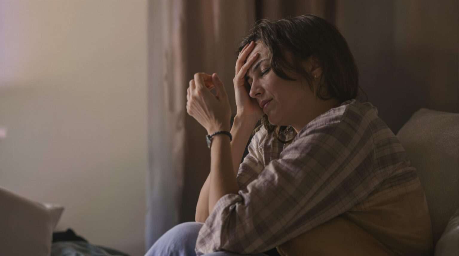 A distressed woman sitting in dim lighting, holding her head in pain, showing Can Hypnosis Help PTSD? How Trauma-Focused Hypnotherapy Creates Lasting Change.