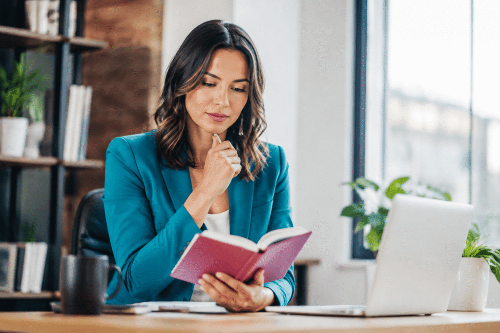 Woman in a teal blazer sits at a desk, reading a pink notebook with a laptop nearby.