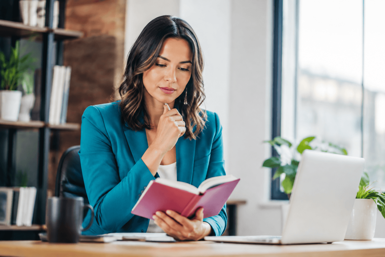 Woman in a teal blazer sits at a desk, reading a pink notebook with a laptop nearby.